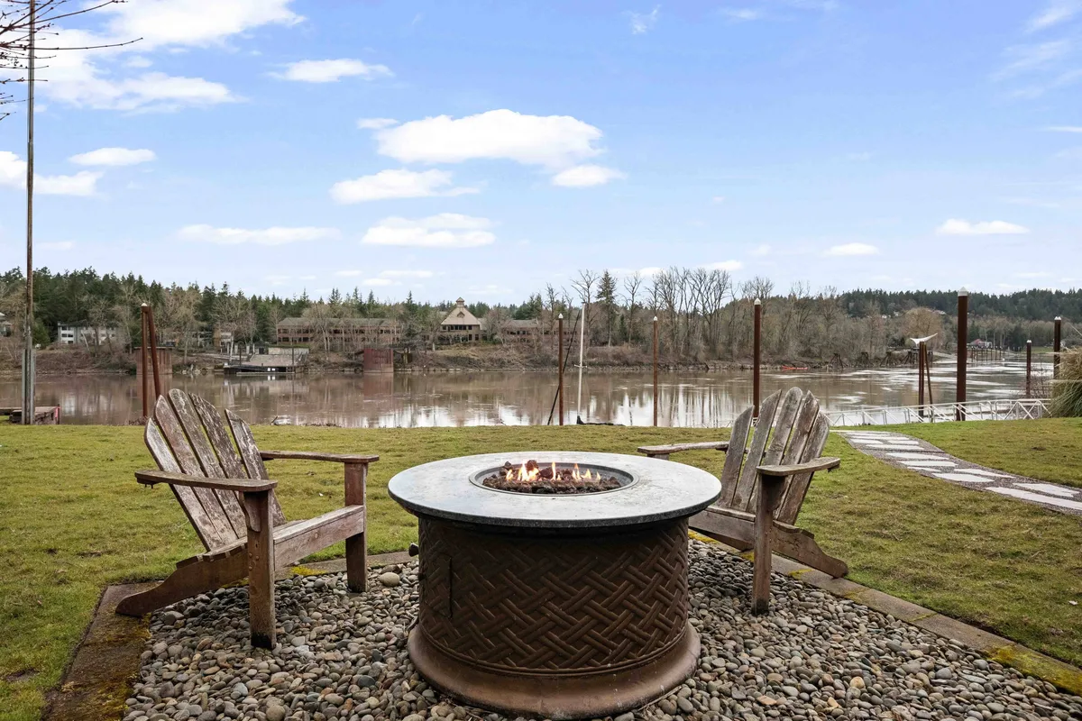 An inviting riverside fire pit area with comfortable chairs arranged in a circle, facing a glowing fire. The river is visible just beyond, providing a serene and scenic backdrop for evening gatherings.