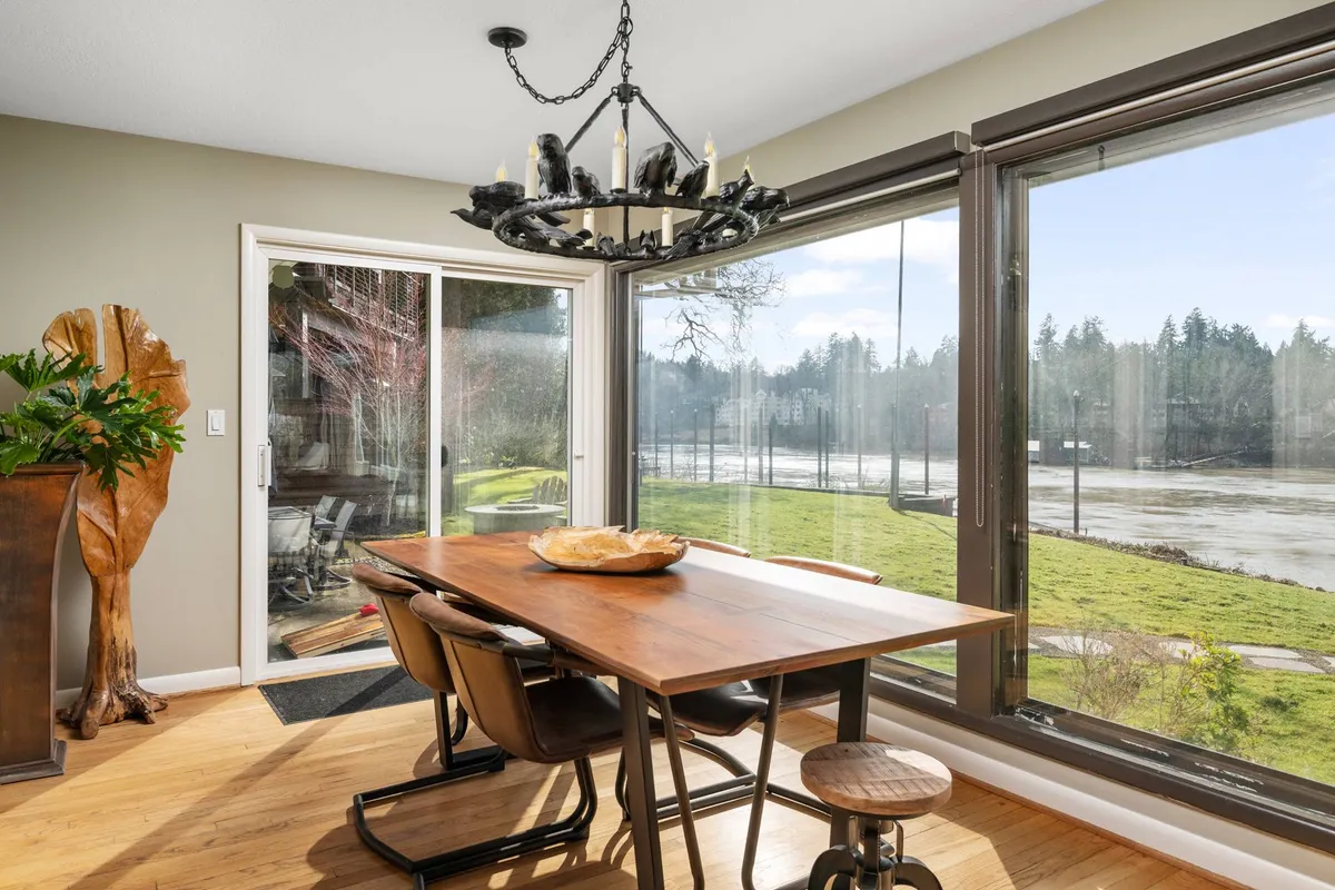 A spacious dining area bathed in natural light, with a chandelier hanging over a large wooden table. Massive windows provide breathtaking views of the river, seamlessly connecting indoor gathering with the riverscape outside. The décor is warm and inviting, ideal for shared family meals or entertaining guests.