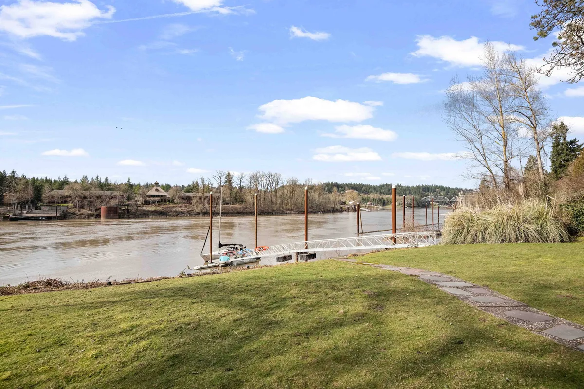 A serene river view with a private dock, lush green grass, and mature trees lining the waterfront. The peaceful water and open sky create a calming backdrop, perfect for soaking in views or enjoying quiet moments with friends.
