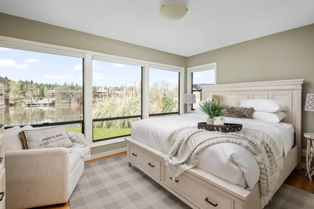 A serene king bedroom featuring large windows that offer stunning views of the water, a cozy bed with plush bedding, an inviting armchair with a decorative pillow, and a side table with a potted plant. Natural light fills the room, creating a peaceful retreat ideal for rest and relaxation.