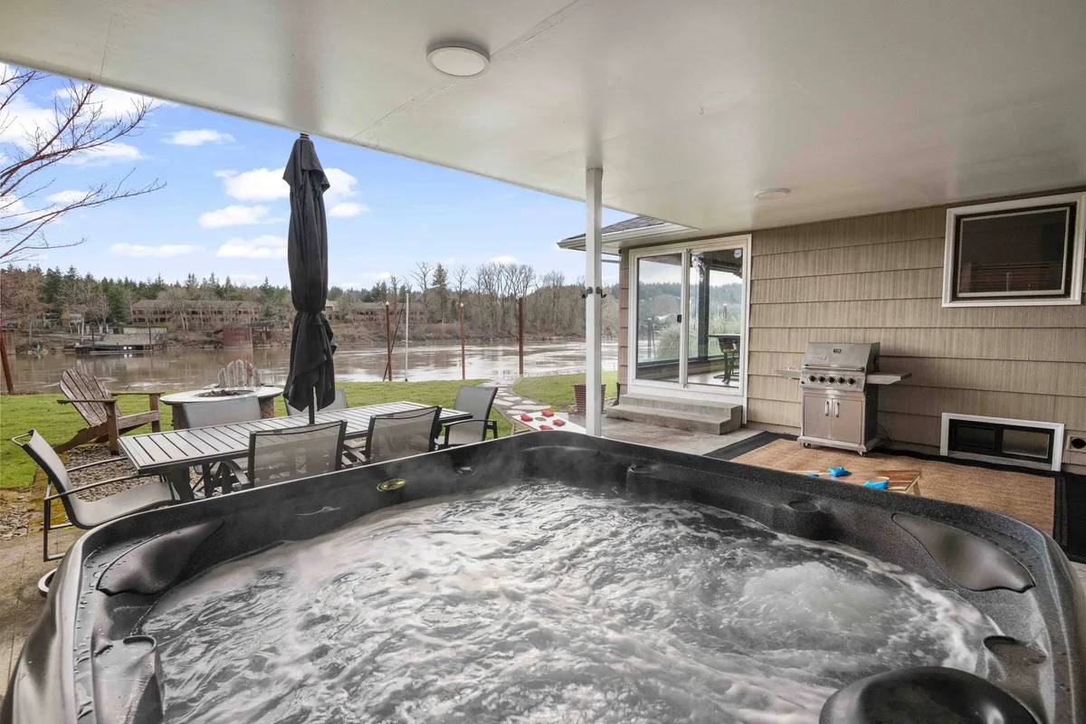This exterior image captures a serene backyard oasis with a hot tub, inviting outdoor seating area, and a panoramic view of the Willamette River. The tranquil setting is perfect for relaxing, family gatherings, or enjoying sunset views from the hot tub.