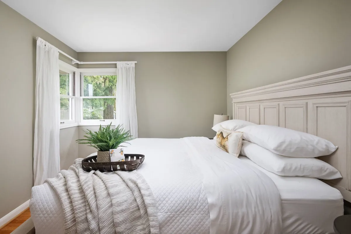 A tastefully decorated king bedroom with a plush bed adorned with decorative pillows, bathed in natural light streaming through a window. A green plant on a tray adds a touch of freshness, and the room exudes a sense of peace and comfort—perfect for winding down or enjoying a quiet morning coffee.
