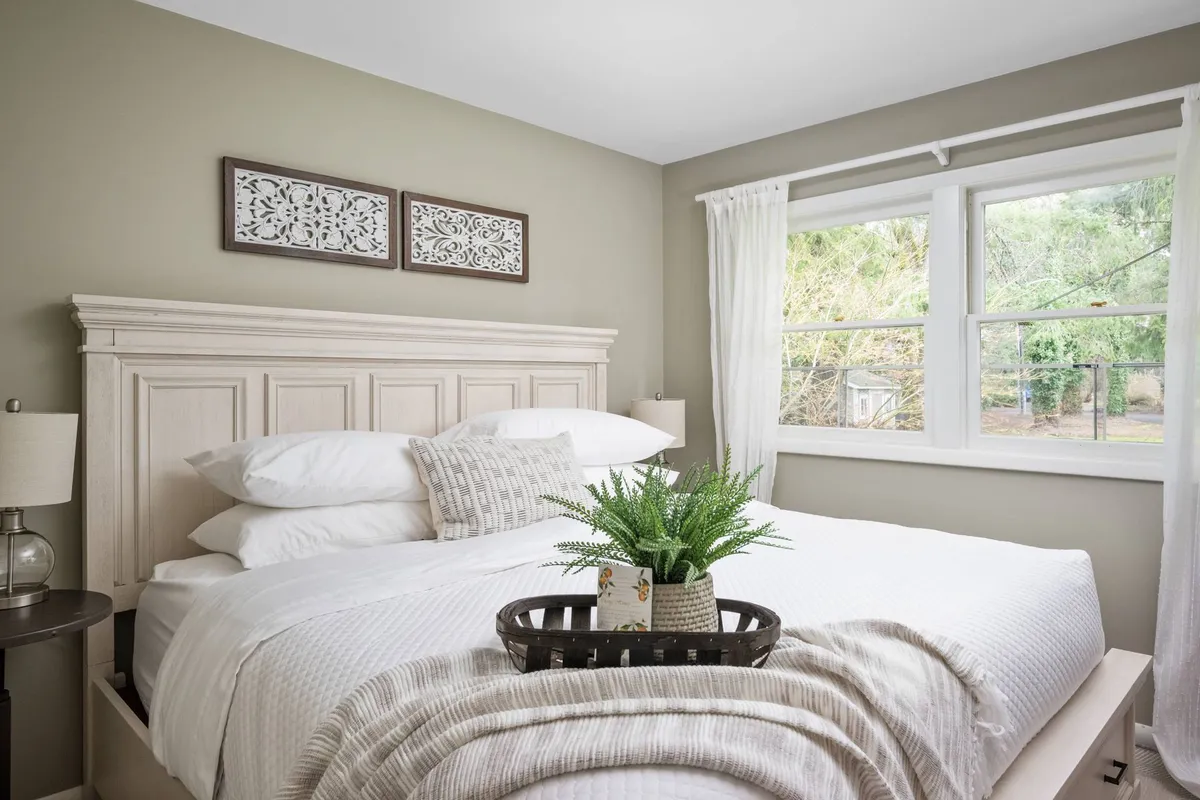 An inviting king bedroom with white linens, decorative wall art, and a green potted plant. The calming color palette and uncluttered design make the room feel peaceful and private, ideal for restful nights or morning relaxation.