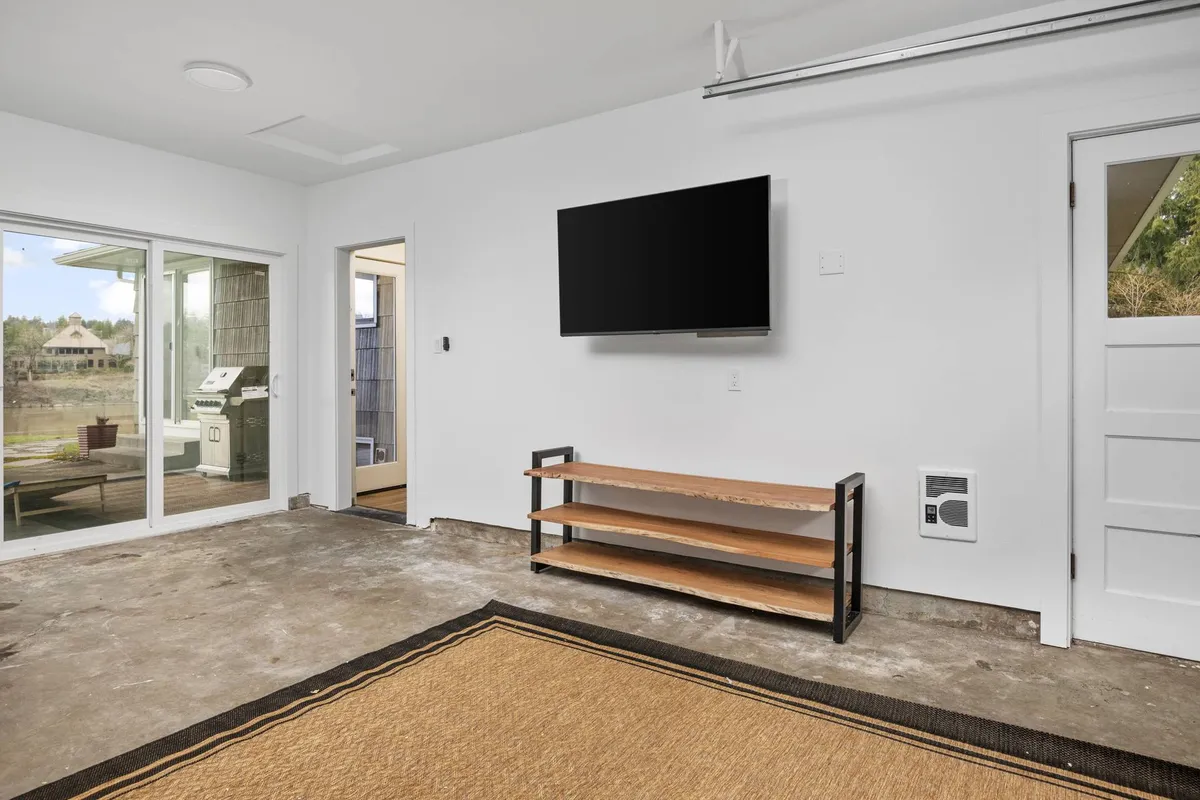 A casual interior scene with a wall-mounted television, a shelving unit for storage, and a large sliding glass door bringing in natural light. The layout suggests a relaxed environment suitable for socializing or cozying up for a movie marathon.