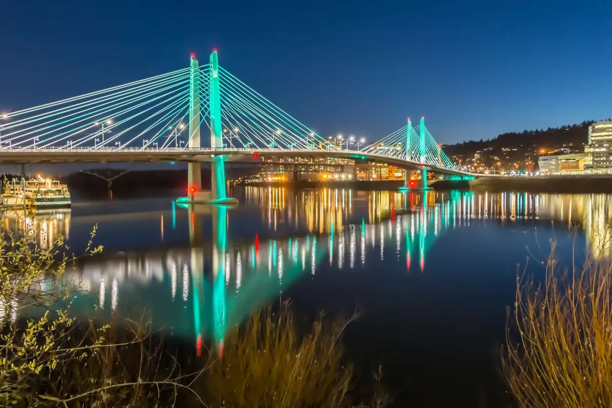A beautifully illuminated bridge reflecting on the water at night, set against the backdrop of city lights. The image conveys both tranquility and vibrant urban energy, hinting at nearby nightlife and dining options in Portland while maintaining a peaceful riverside ambiance.