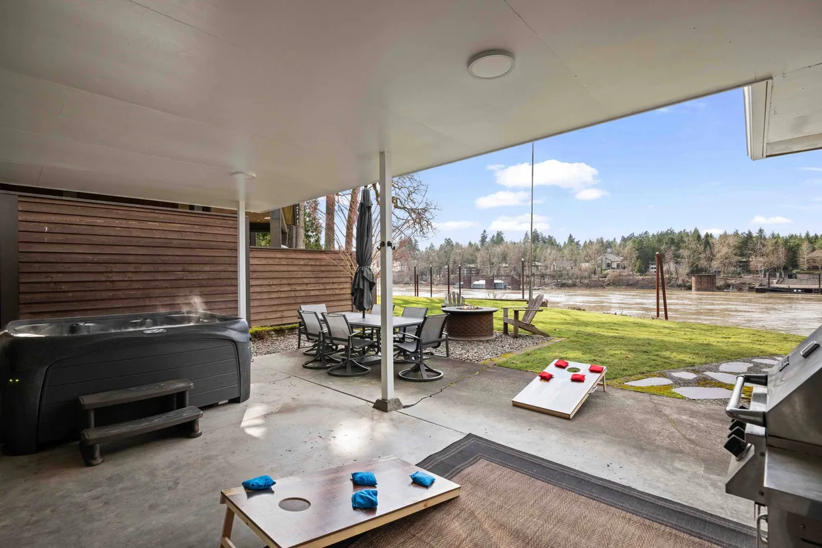 A lively backyard scene featuring a bubbling hot tub, an outdoor dining table set for a meal, a cornhole game on the expansive green lawn, and a grill—all with a tranquil lake view in the background. The atmosphere is relaxed and inviting, hinting at both recreation and relaxation.