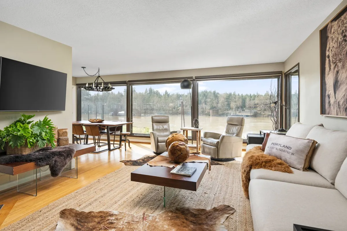 The image showcases a sunlit, open-concept living and dining area with large windows offering tranquil views of riverfront nature. A modern, beige sofa and elegant dining set create an inviting space for relaxation or gathering with loved ones, complemented by lush greenery and curated artwork.